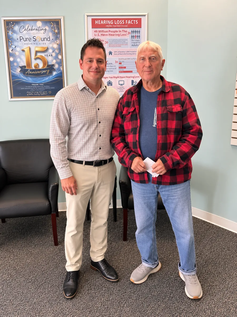 Two men standing in the Pure Sound Hearing Aids office; older man in a red plaid shirt holding an envelope, with Hearing Instrument Specialist Sean Mark beside him.