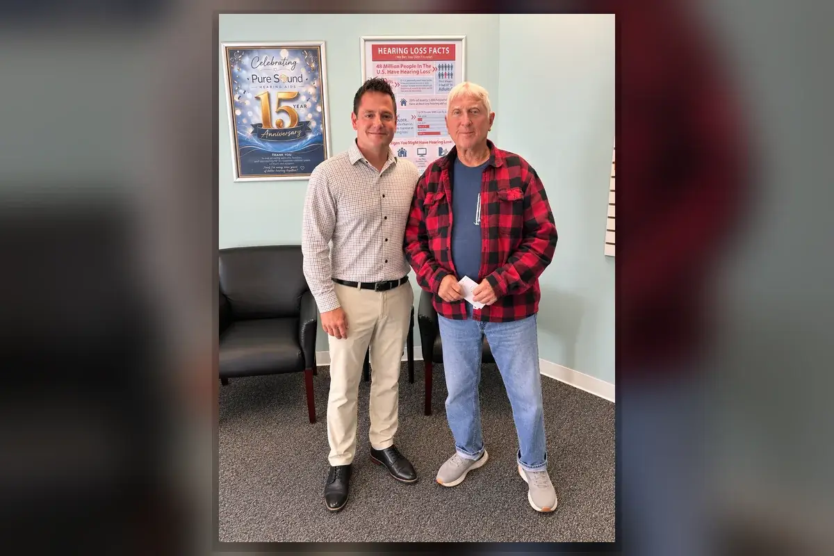 Two men standing in the Pure Sound Hearing Aids office; older man in a red plaid shirt holding an envelope, with Hearing Instrument Specialist Sean Mark beside him.